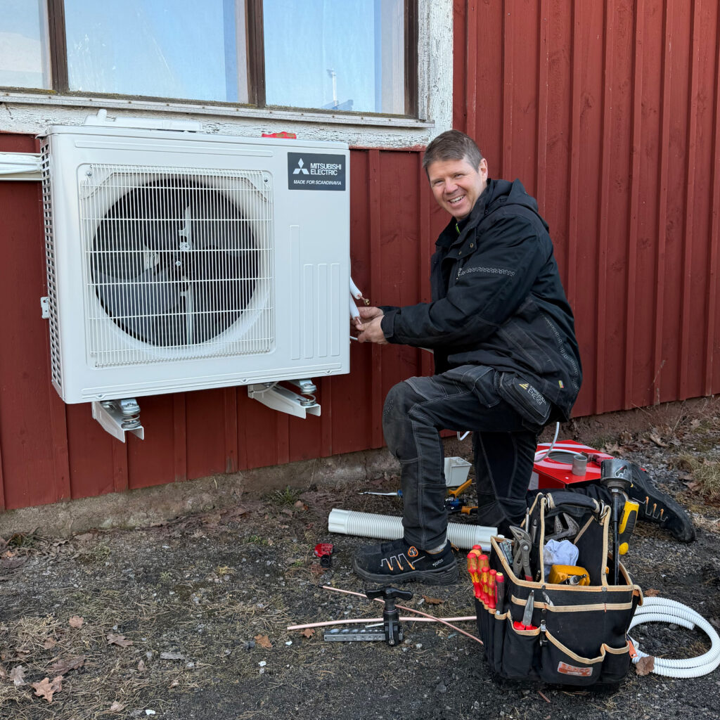Magnus installerar en luftvärmepump på en röd träfasad.