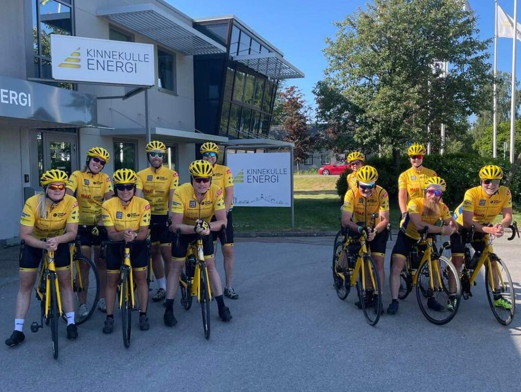 11 cyklister från Team Rynkeby Skaraborg som lutar sig över deras landsvägscyklar. alla har hjälp och samma gula kläder. De står framför Kinnekulle Energis lokaler.