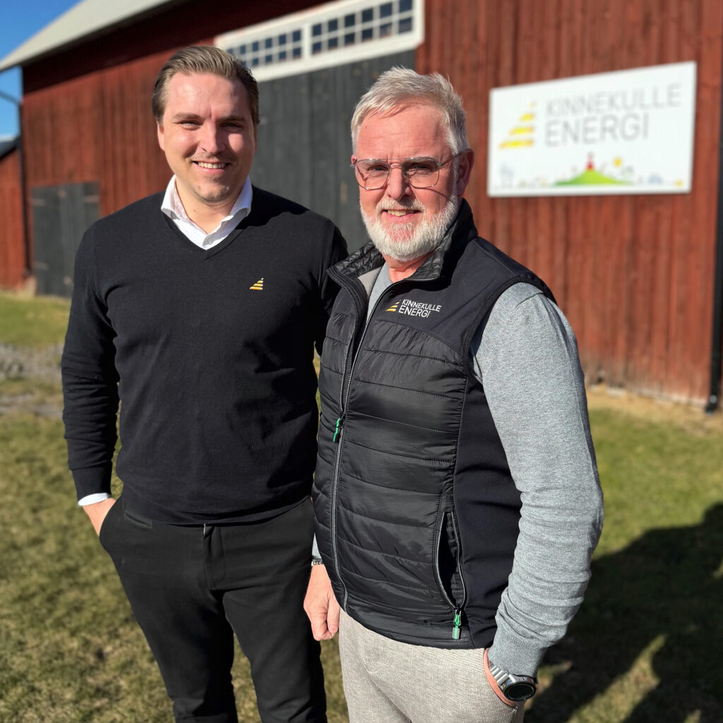 två män som står ute i solen framför en ladugård.